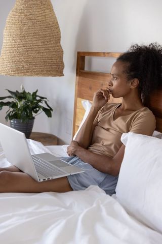 African american woman working on laptop in cozy bedroom, thoughtful remote work
