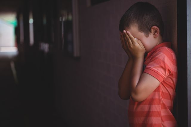 Young boy standing in a school corridor, covering his face with his hands, appearing sad and upset. This image can be used to depict themes of childhood stress, bullying, loneliness, and mental health issues in educational settings. Ideal for articles, blogs, and campaigns focused on child welfare, education, and emotional well-being.