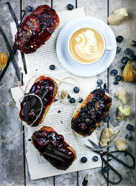 Showing assortment of freshly baked pastries spread with jam next to cappuccino on rustic wooden table. Background includes scattered blueberries, ginger pieces, and vintage scissors. Perfect for culinary blogs, breakfast menus, coffee shop promotions, or baking classes.