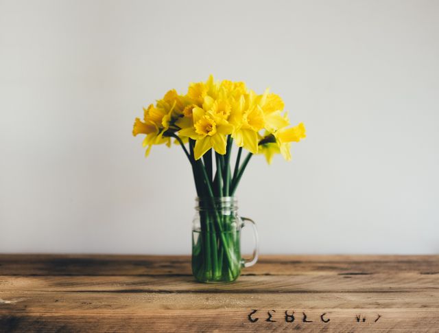 Bright bouquet of yellow daffodils placed in a clear mason jar on a wooden table against a plain background. Ideal for use in springtime-themed projects, articles about home decor, nature, or floral arrangements. Suitable for blogs, social media posts, or print materials focusing on interior design or gardening.