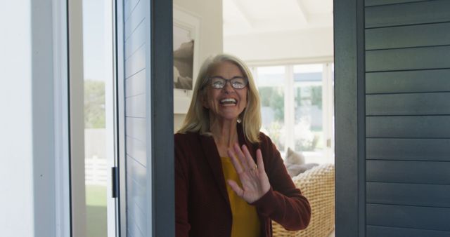 Senior woman stands at front door, waving and smiling warmly. Her bright and modern living room is visible in the background, suggesting a welcoming and hospitable environment. Suitable for themes related to home, aging, hospitality, family visits, and senior lifestyle.
