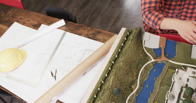 Architect's hands, visible from the shoulders and dressed in a plaid shirt, working with a landscape model and blueprints on a wooden table. The scene also includes a yellow helmet and technical drawings. Useful for themes involving architecture, engineering, planning, or project design.