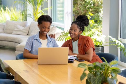 African mother and teenage daughter studying on laptop and smiling in bright modern home