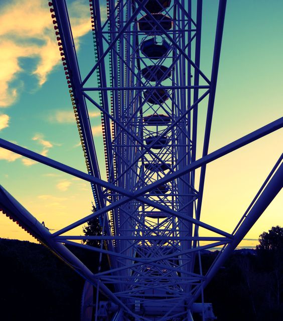 This image captures a detailed view of a Ferris wheel's metal structure as a silhouette against the backdrop of a sunset sky. The geometric design elements emphasize the engineering and architectural beauty while the vibrant colors of the sunset enhance the visual appeal. Ideal for use in travel brochures, themes on adventure, amusement park promotions, and architectural magazines.
