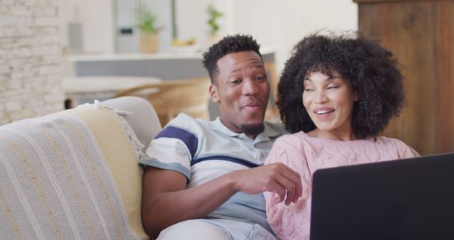 Happy African American couple sitting on sofa at home, enjoying time together while using laptop. Ideal for advertisements related to technology services, family bonding, streaming services, and home lifestyle. Great for blogs or articles focusing on modern living, relaxation methods, and digital age connectivity.