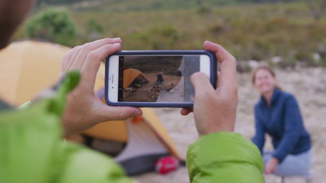 Joyful couple on a mountain camping trip, with the woman cooking sausages over campfire and the man capturing moment with smartphone. Ideal for travel blogs, outdoor adventure promotions, or advertising camping equipment.