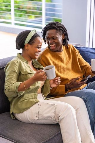 Laughing african american couple sharing coffee on modern sofa by sunny balcony