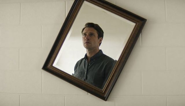 Man reflecting in tilted vintage mirror against painted block wall, introspective mood