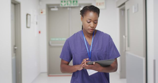 African-American female nurse wearing blue scrubs walking in hospital corridor and using tablet. This image is useful for healthcare technology, hospital management, medical professional websites, nursing blogs, and educational materials related to healthcare practices and technology integration.