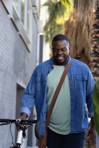 African american man walking bicycle through sunny urban alley carrying messenger bag