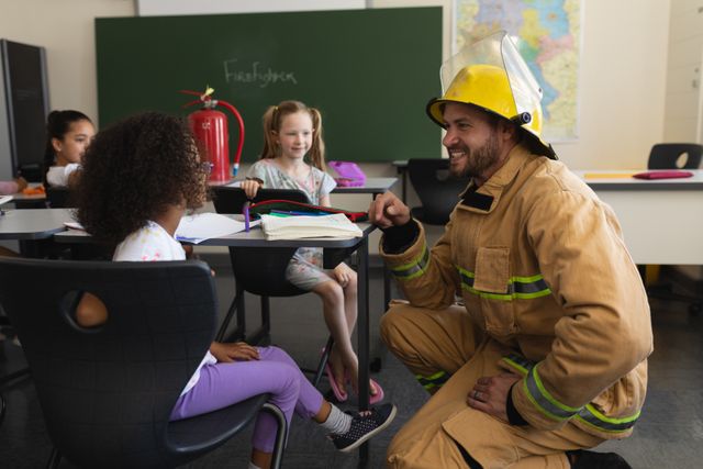 Side view of male Caucasian firefighter on his knees teaching schoolgirls about fire safety with smile in classroom of elementary school