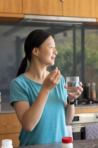 Mature asian woman taking pill with glass of water in modern kitchen