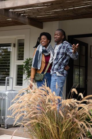 Couple greeted on porch with luggage under pergola