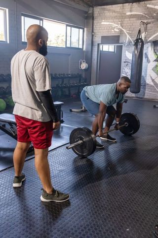 African man deadlifting while trainer watches in industrial gym with mural and daylight