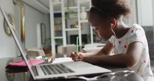 Young black girl wearing earphones, concentrating on homework while using laptop. Light-filled modern home interior in background. Could be used for illustrating remote learning, online education platforms, or children’s educational materials.