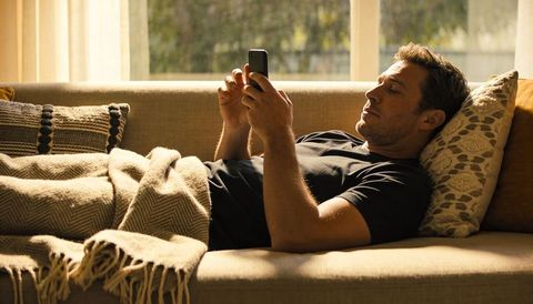Man relaxing on sofa scrolling smartphone under blanket in sunlit living room