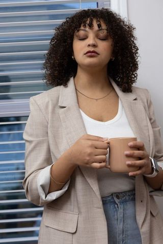 Contemplative professional woman holding beige mug by window blinds, morning coffee break