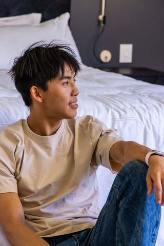 Asian man sitting on floor leaning against bed in modern bedroom wearing smartwatch casual vibe