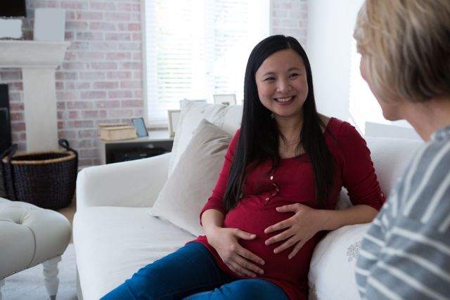 Pregnant woman sitting on a couch, smiling and chatting with a friend in a cozy living room. Ideal for use in articles or advertisements related to pregnancy, maternity, friendship, and home life. Suitable for blogs, social media posts, and parenting websites.