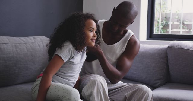 A father is styling his young daughter's curly hair in their living room. This image captures a tender moment of parent-child bonding, emphasizing caring and familial love. Suitable for use in parenting articles, family-oriented advertisements, or blogs about fatherhood and family relationships.