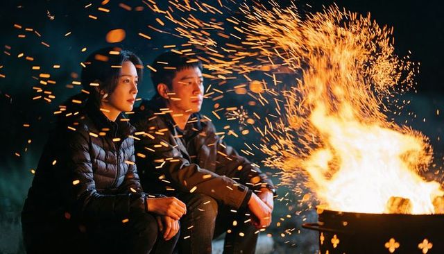 Couple sitting beside blazing metal fire barrel warming hands, sparks flying night romance