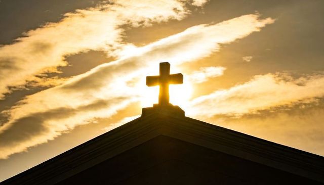 Backlit church cross silhouette at golden sunrise with radiant halo clouds
