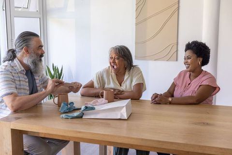 Multiracial family sharing card and coffee, senior woman laughing at wooden dining table