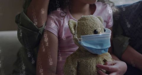 Child cuddling teddy wearing surgical mask on sofa with caregiver and binary overlay
