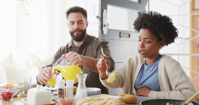 Image of happy diverse couple eating and pouring juice at breakfast table. Happiness, inclusivity, free time, togetherness and domestic life.
