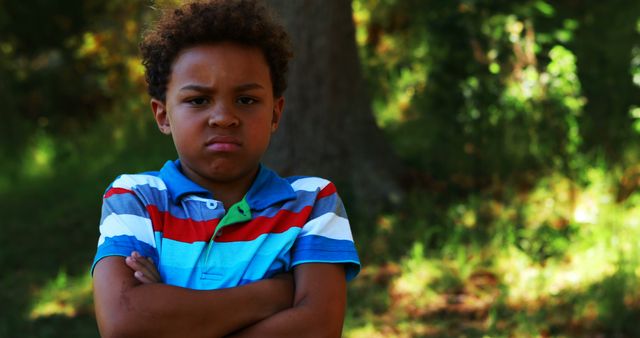 A young African American boy appears upset with his arms crossed, standing outdoors with trees in the background, with copy space. His expression and body language suggest he might be feeling frustrated or defiant.