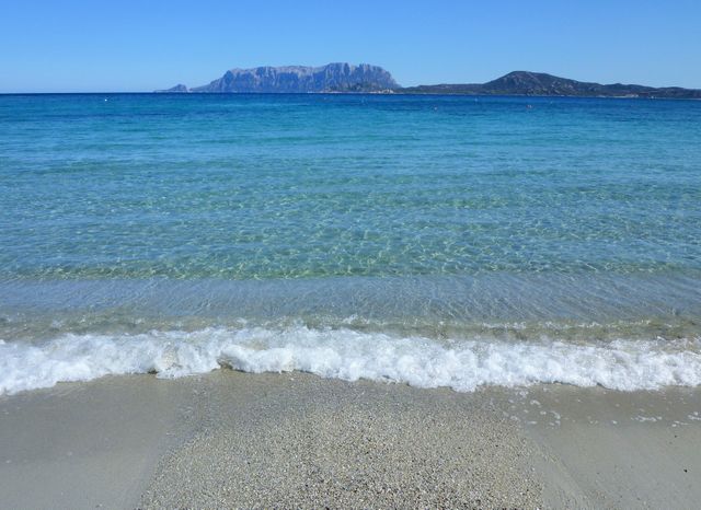 This image shows a tranquil beach with turquoise waters and a distant island. The waves gently lap the sandy shore under a clear blue sky, with mountainous terrain visible in the background. Ideal for travel brochures, vacation advertisements, and websites promoting beach destinations. The peaceful setting can also be used for wellness and relaxation themes.