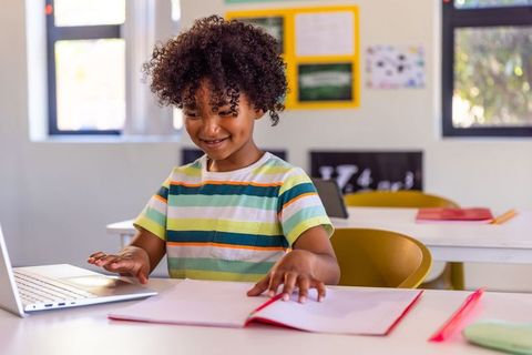 Smiling african american boy using laptop and taking notes in bright elementary classroom