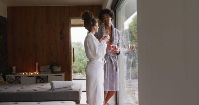 Image of relaxed diverse couple in bath robes drinking tea and talking at health spa. Vacation, togetherness, relaxation, health and inclusivity concept.