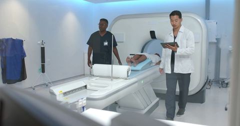 Diverse medical team positioning patient on ct scanner while doctor reviewing tablet in radiology su