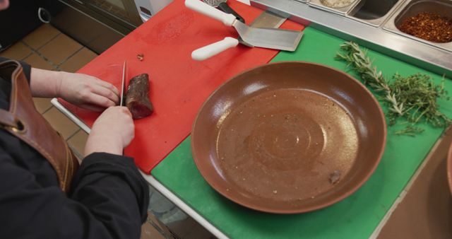 Professional chef slicing meat on chopping board in kitchen. Culinary tools, herbs, and large brown plate visible. Perfect for food blogs, restaurant promotions, culinary school materials.