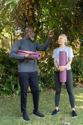 Diverse senior couple holding purple yoga mats preparing outdoor yoga beneath fruiting tree