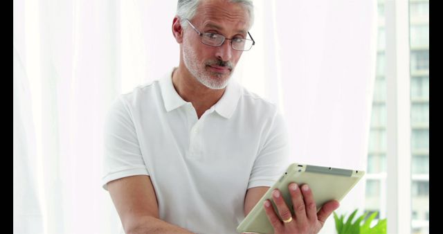 An elderly man with silver hair stands indoors wearing a white shirt and glasses while engaging with a tablet and displaying a skeptical expression. This image is ideal for concepts related to skepticism, analysis, technology use among seniors, internet browsing, and modern lifestyle.