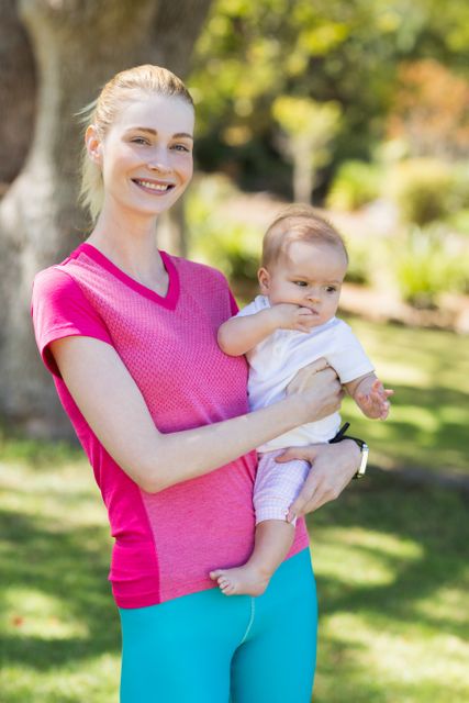 Smiling mother holding her baby in a park, enjoying a sunny day. Ideal for use in parenting blogs, family-oriented advertisements, and promotional materials for outdoor activities or family products.