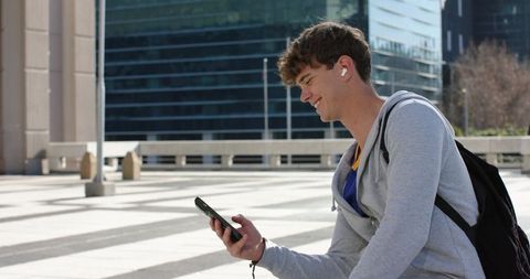 Young man sitting on urban plaza ledge checking smartphone, wearing earbuds and backpack