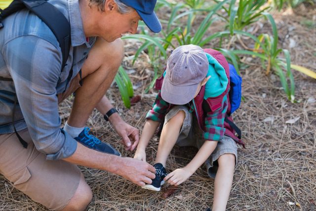 Father tying son's shoelace during a forest hike, showcasing family bonding and outdoor adventure. Ideal for use in parenting blogs, outdoor activity promotions, family-oriented advertisements, and nature exploration articles.