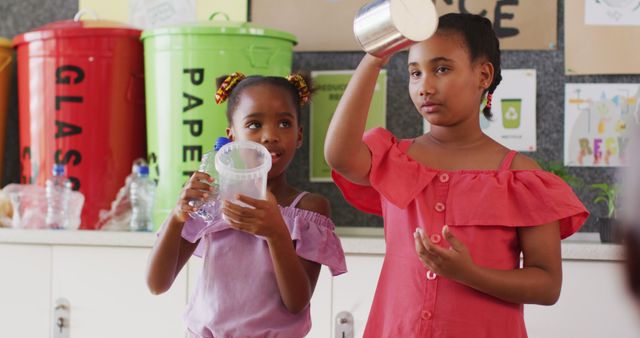 Two young African girls warmly engaging in an eco-friendly lesson, clearly focused on recycling and sustainability. Brightly colored bins for glass and paper are visible in the background, emphasizing environmental responsibility. Ideal for use in educational content, environmental campaigns, and community outreach programs promoting waste management and recycling efforts.
