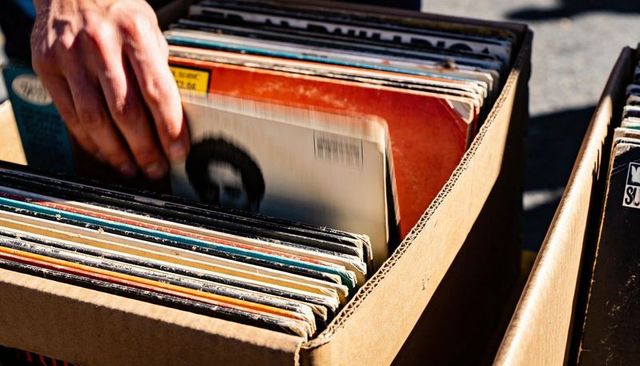 Hand browsing vintage vinyl records in sunlit cardboard box at sidewalk flea market