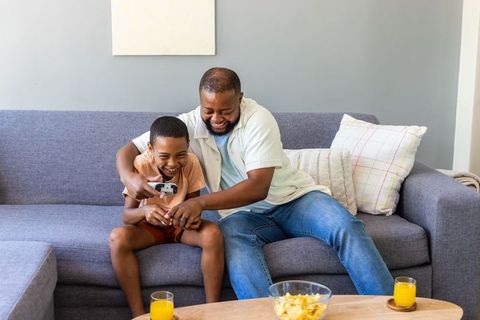 African american father and son laughing while playing video game on gray sofa