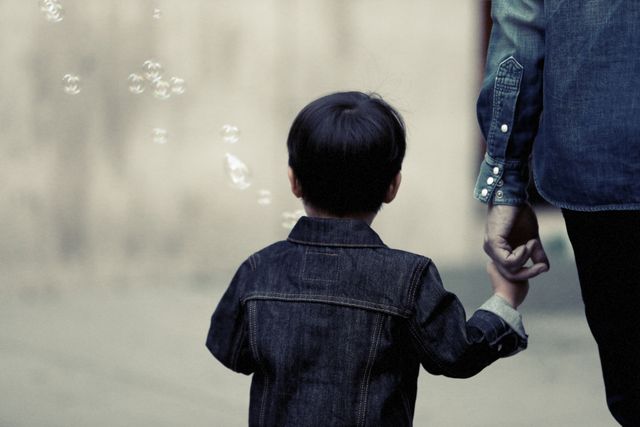 Child holding an adult's hand while walking outdoors, wearing denim jackets. Bubbles are floating in the background. Ideal for themes related to family bonding, parent-child relationships, outdoor activities, trust, and childhood memories.