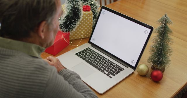 Middle-aged man engaging in a video call on his laptop during Christmas, surrounded by festive decorations such as a small Christmas tree, ornaments, and wrapped gifts. Ideal for concepts related to holiday season communication, virtual celebrations, and staying connected with loved ones during festive times. Perfect for use in marketing materials, holiday greetings, or articles about technology and family interactions.