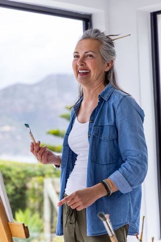 Senior woman painting in sunlit coastal studio smiling while holding paintbrush