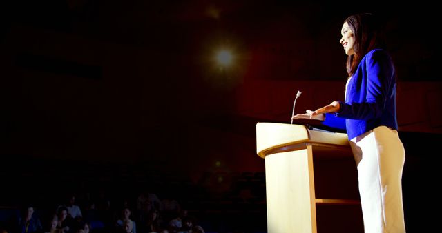 Woman standing at podium delivering influential speech to audience in large auditorium. Ideal for themes related to leadership, business presentations, public speaking, motivational events, and professional development.