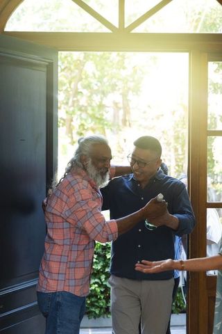 Senior friends embracing and reuniting at sunlit front door holding bottle, welcome home