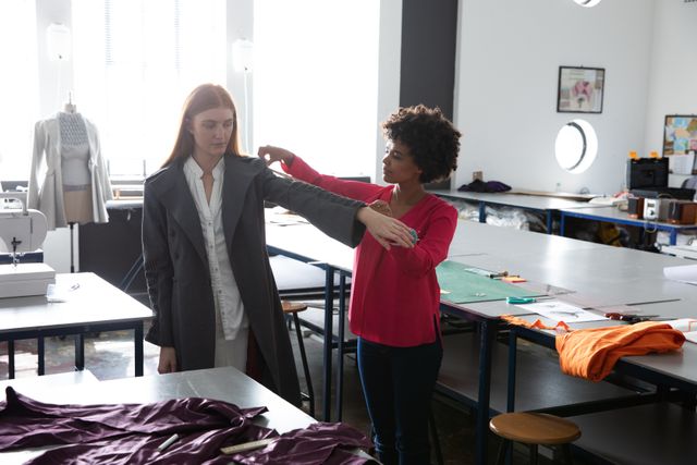 Front view of a young biracial female fashion student measuring a jacket worn by a young Caucasian female student with long red hair, in a studio at fashion college