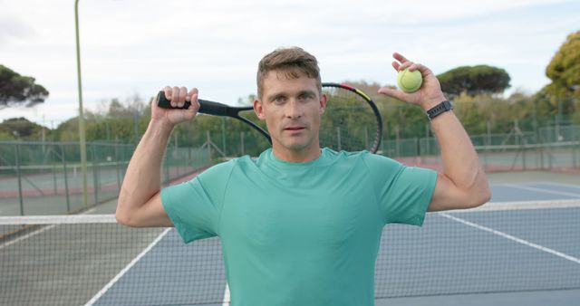 Adult male tennis player standing on an outdoor court, holding a tennis racket over his shoulder and a tennis ball in his other hand. He is wearing a green shirt and appears to be ready for a match. This image could be used for promoting tennis equipment, sportswear, or events, as well as fitness and health campaigns.